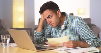 A frustrated man sitting at his laptop with a handful of denied insurance paperwork